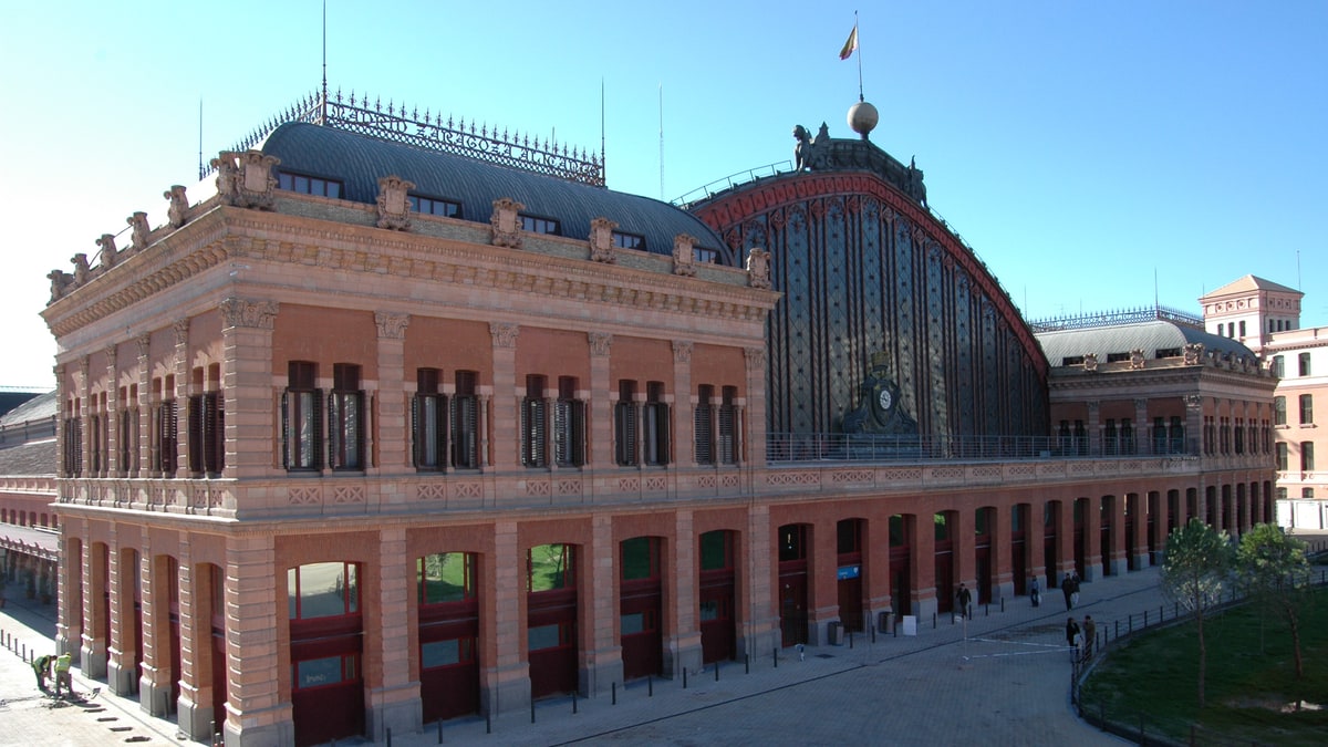 Fachada de ladrillo de la estación de Atocha con viajeros caminando en la explanada