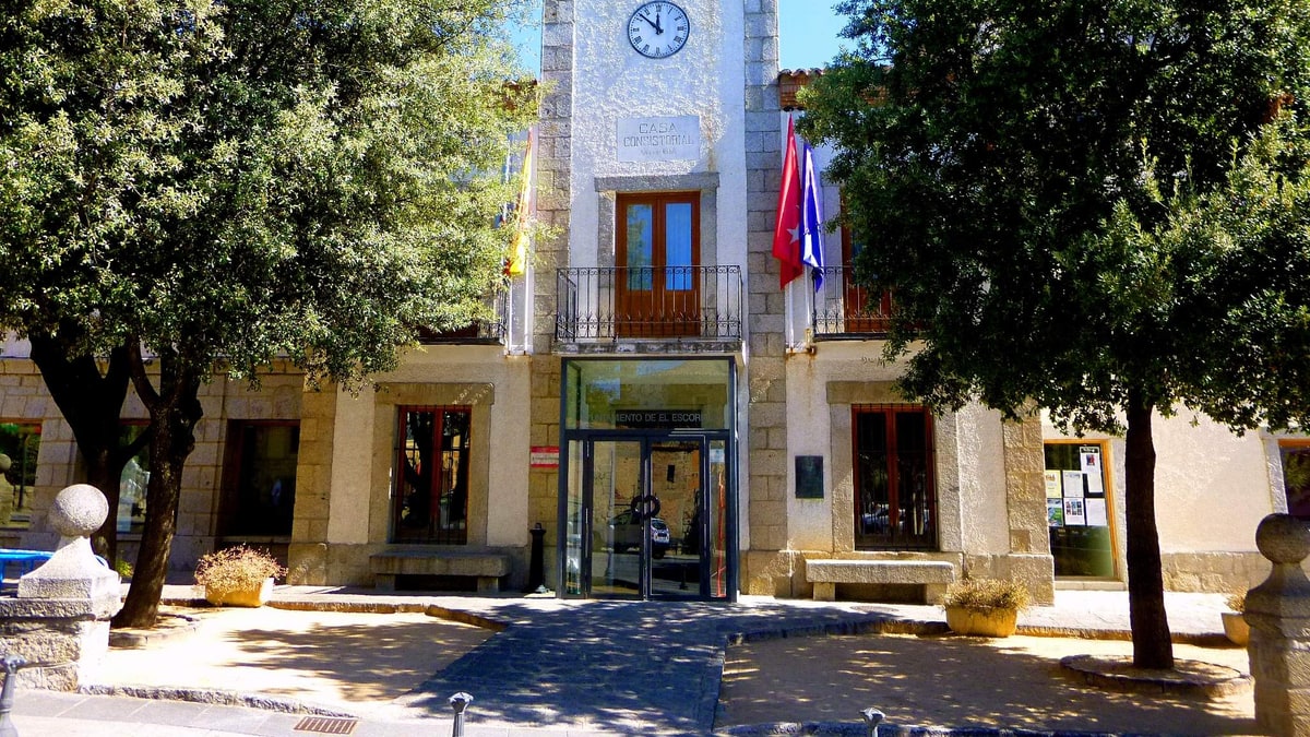 Fachada del Ayuntamiento de El Escorial con reloj, bandera y árboles en la plaza