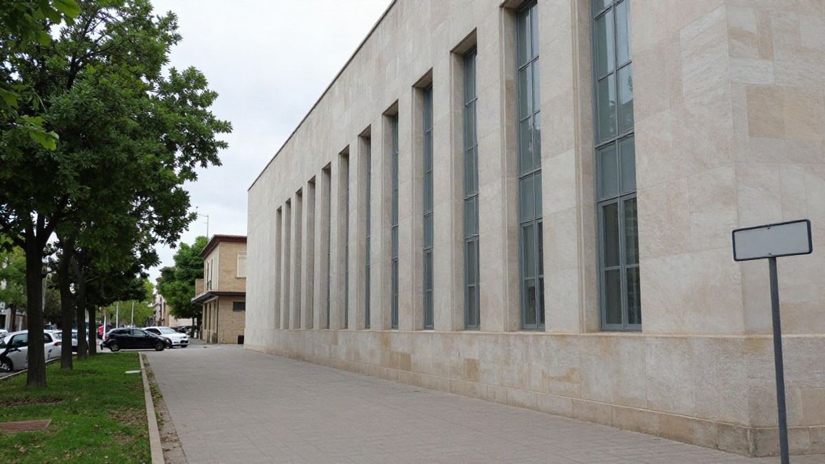 Illustrative image: sober facade of an institutional building next to a tree-lined walkway, under a cloudy daytime sky.