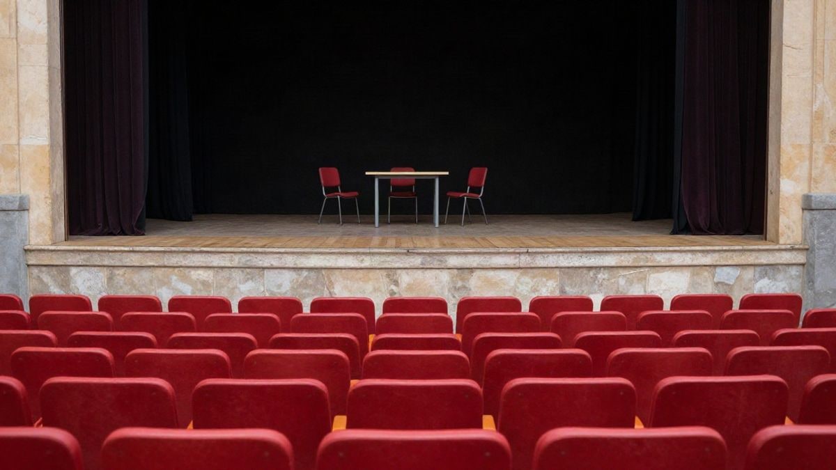Imagen ilustrativa: butacas rojas de un teatro vacío frente a un escenario con sillas y una mesa